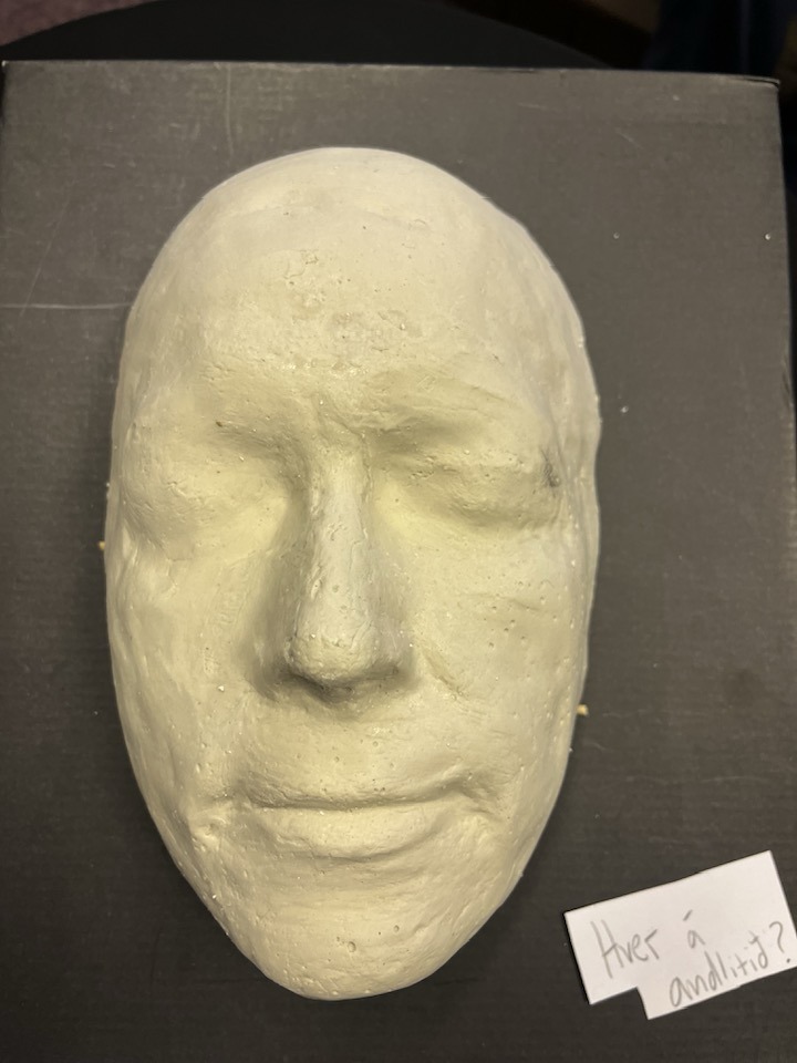 Afsteypa af andliti Siggu komin upp á vegg. Fyrir neðan er miði sem á stendur: Hver skildi þetta vera.