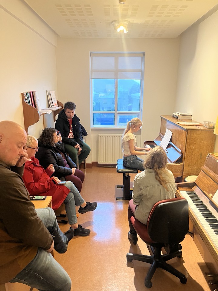 Gestir á opnu húsi fylgjast með nemanda spila á píanó inni í kennslustofu.