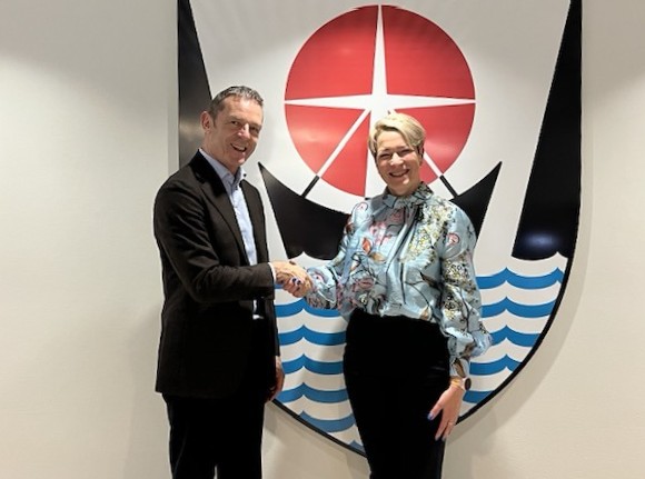 Danski sendiherrann og Sigríður takast í hendur fyrir framan merki Ísafjarðarbæjar á vegg í afgreiðslu sveitarfélagsins.