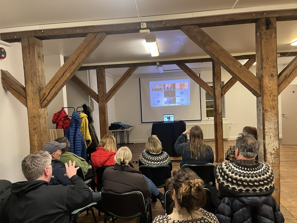 Séð aftan á fundargesti í Rögnvaldarsal í Edinborgarhúsinu. Aftast á mynd er skjávarpatjald þar sem sjást gestir sem eru viðstaddir í gegnum fjarfundabúnað.