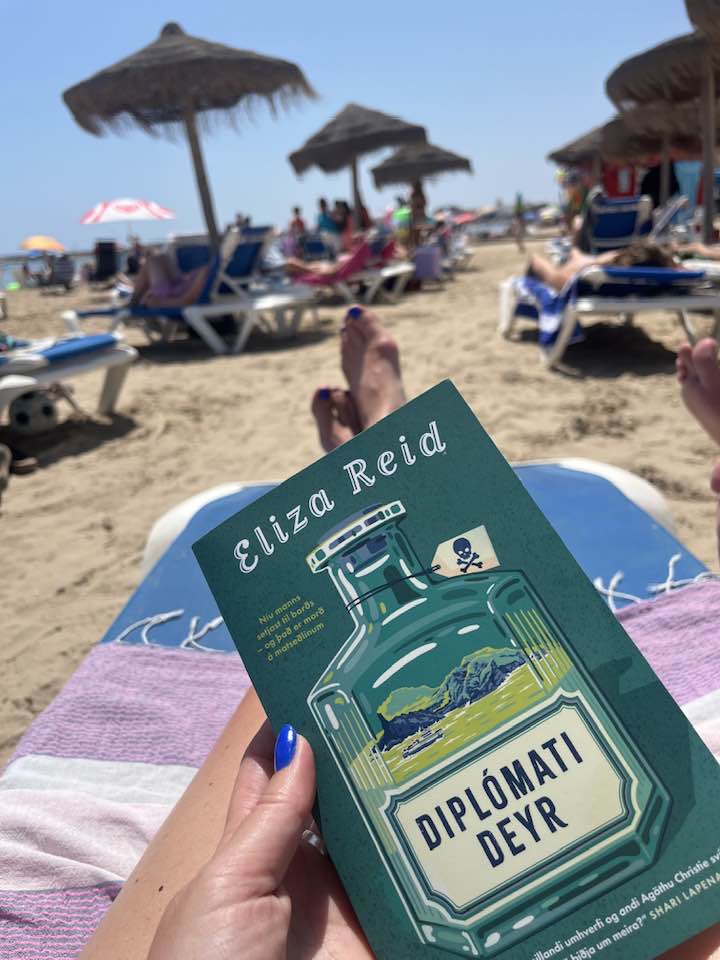 Mynd tekin af sólbekk á strönd. Sigga heldur í bókina Diplómati deyr. Í bakgrunni má sjá fólk á sólbekkjum, undir sólhlífum.