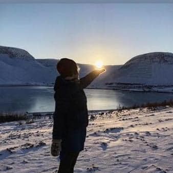 Kona stendur úti í Skutulsfirði, fjöll og sól að setjast í bakgrunni. Hún heldur handleggnum út þannig að það er eins og sólin sé í lófa hennar.
