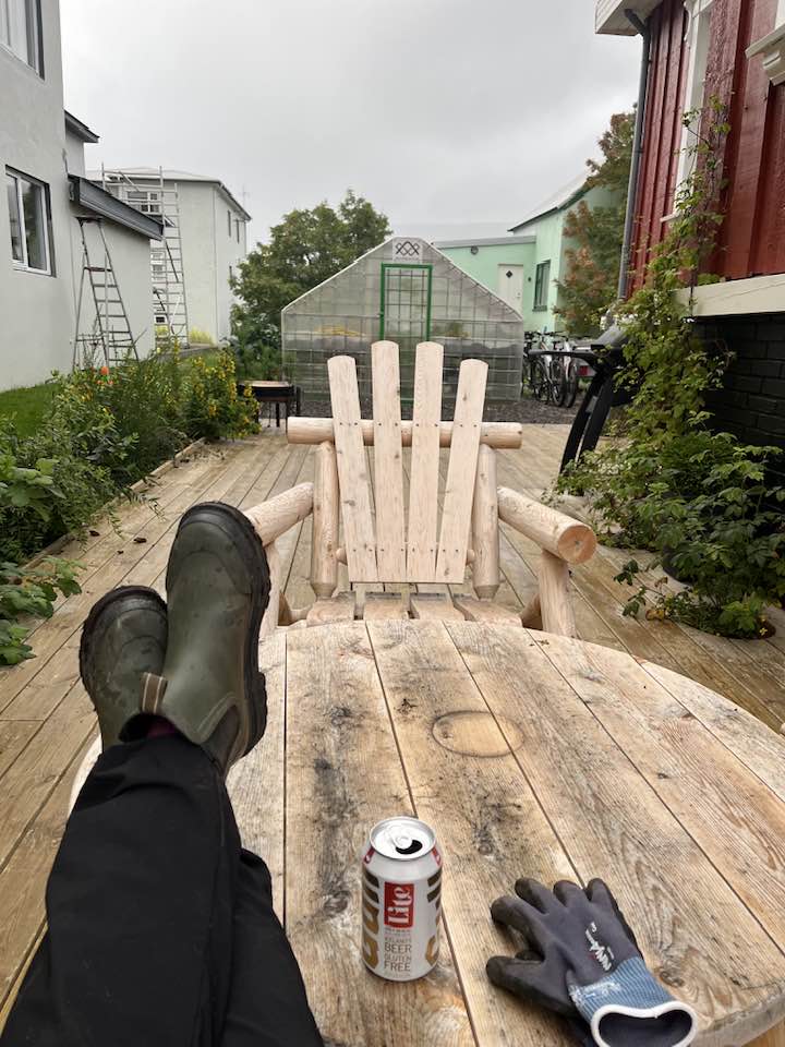 Mynd tekin úr garðstól á palli við hús Siggu á Suðureyri. Fætur uppi á borði, bjór og vinnuhanskar í forgrunni. Pallur, garður og gróðurhús í bakgrunni