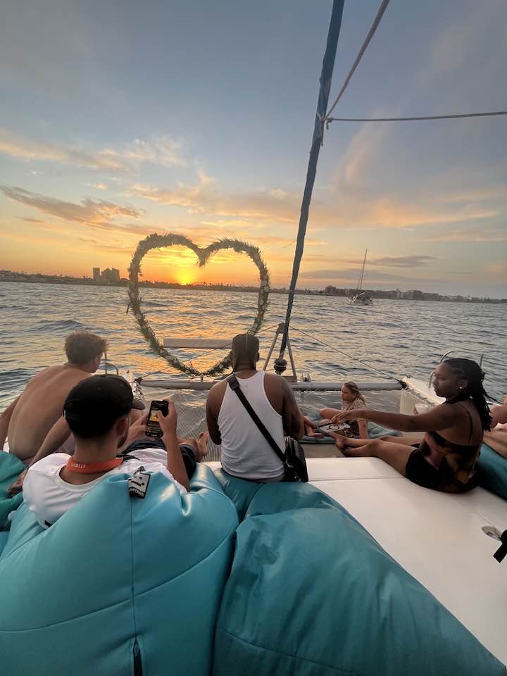 Torrevieja séð frá sjó, á skútu. Farþegar horfa á sólarlagið. Á skut skútunnar er stór blómakrans sem er í laginu eins og hjarta.