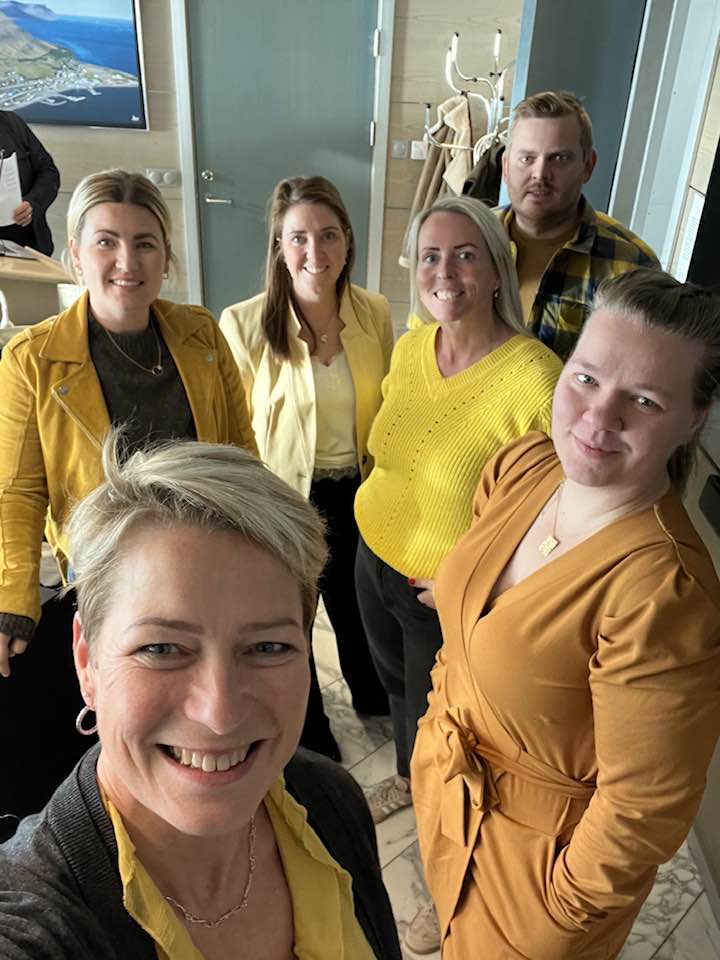 Hópmynd af fimm fulltrúum í bæjarstjórn auk bæjarritara í gulum fötum. Myndin er tekin í fundarsal bæjarstjórnar og hópurinn stendur þétt saman og horfir upp í myndavélina.