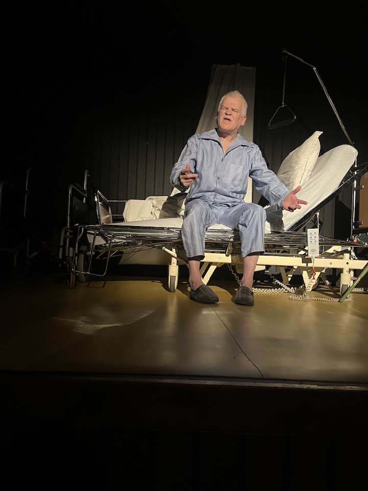 Elfar Logi á sviði í Kómedíuleikhúsinu. Hann situr í sjúkrarúmi og er klæddur náttfötum.