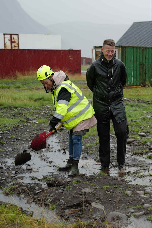 Sigga tekur fyrstu skóflustunguna með rauðri stunguskóflu. Gylfi formaður bæjarráðs stendur hjá.